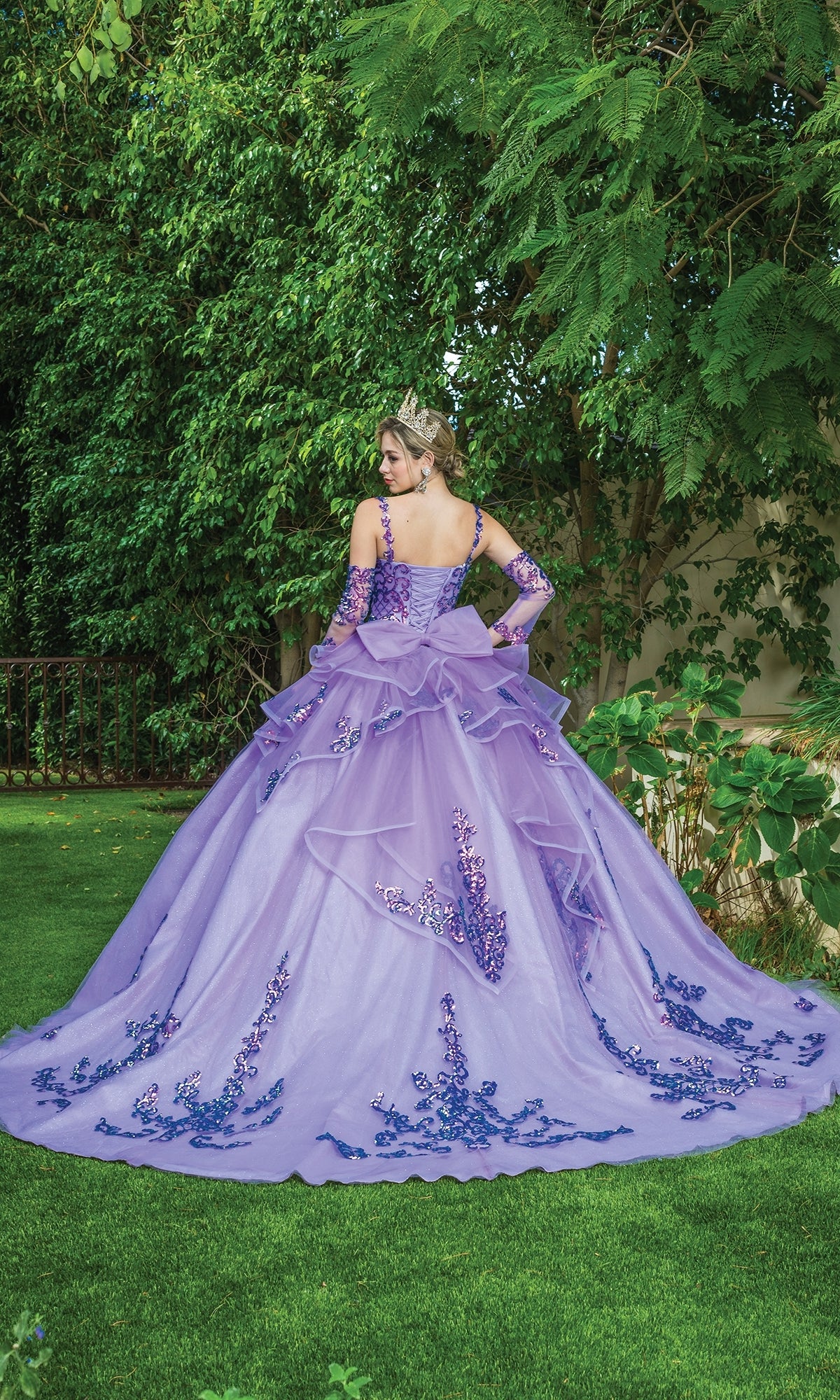 A person wears the Quinceanera Dress 1652 By Dancing Queen, a purple a-line ball gown with blue floral embroidery and a crown, standing on grass among green trees and plants, facing away.